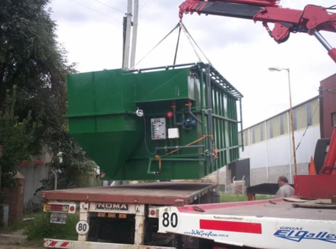 Equipo compacto verde izado con una grúa sobre un camión, listo para su instalación en obra